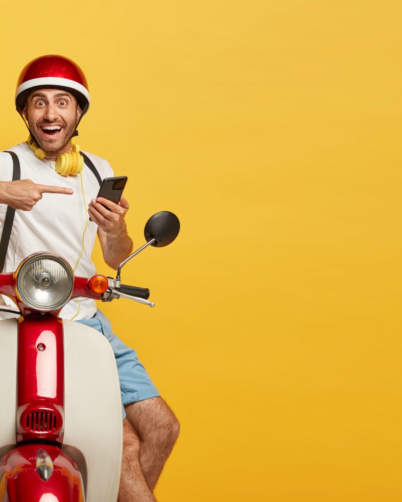 Isolated shot of happy delivery man points at smartphone used for finding route or right address, contacts with customer, poses with rucksack on motorbike, blank copy space on yellow background